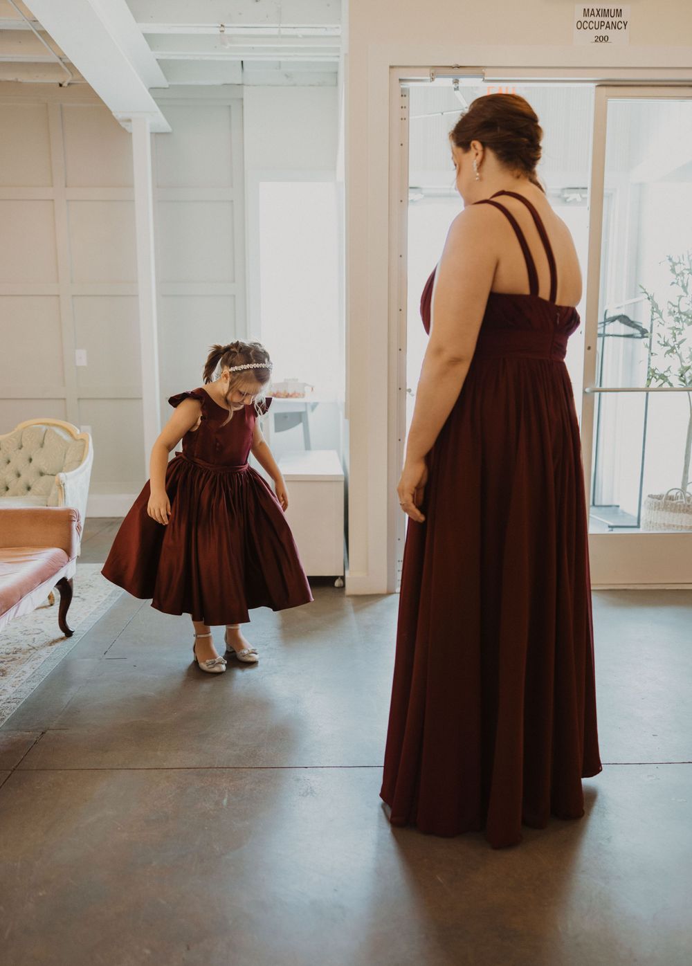 Bridesmaid and flower girl in matching burgundy dresses standing in venue room.
