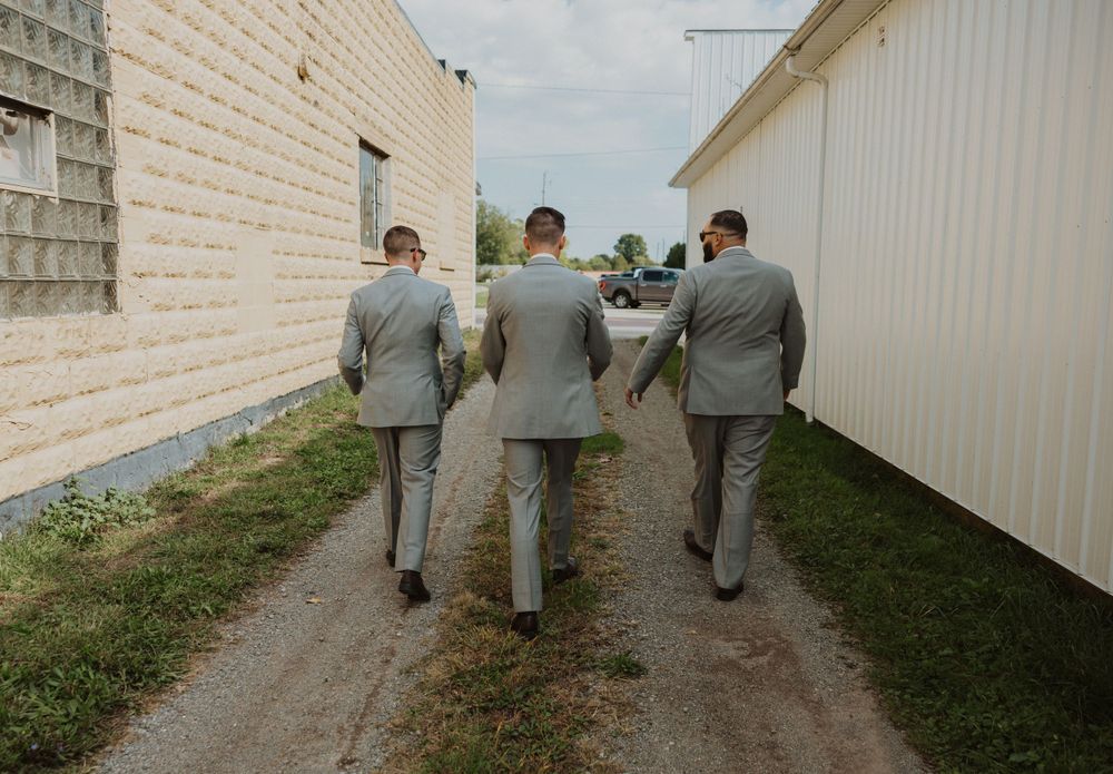 Groomsmen in light gray suits walking along white building exterior.