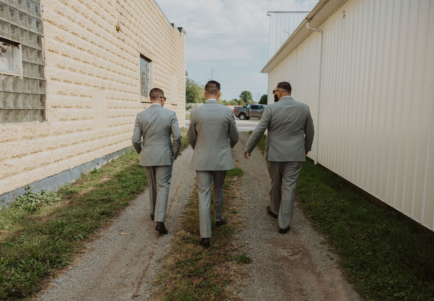 Groomsmen in light gray suits walking along white building exterior.