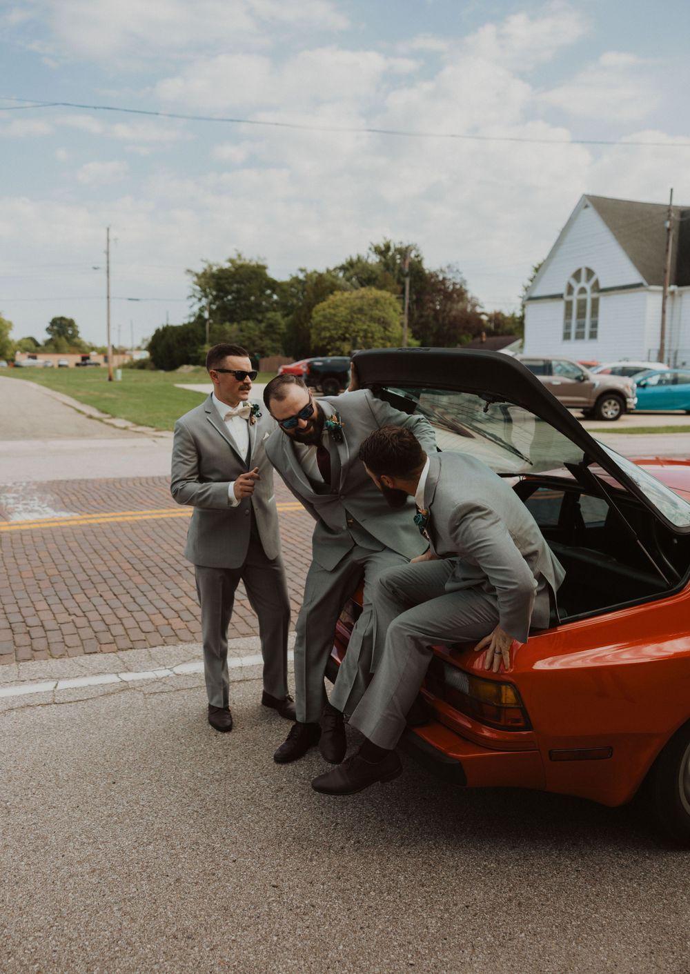 Groomsmen in gray suits examining classic Ferrari sports car.