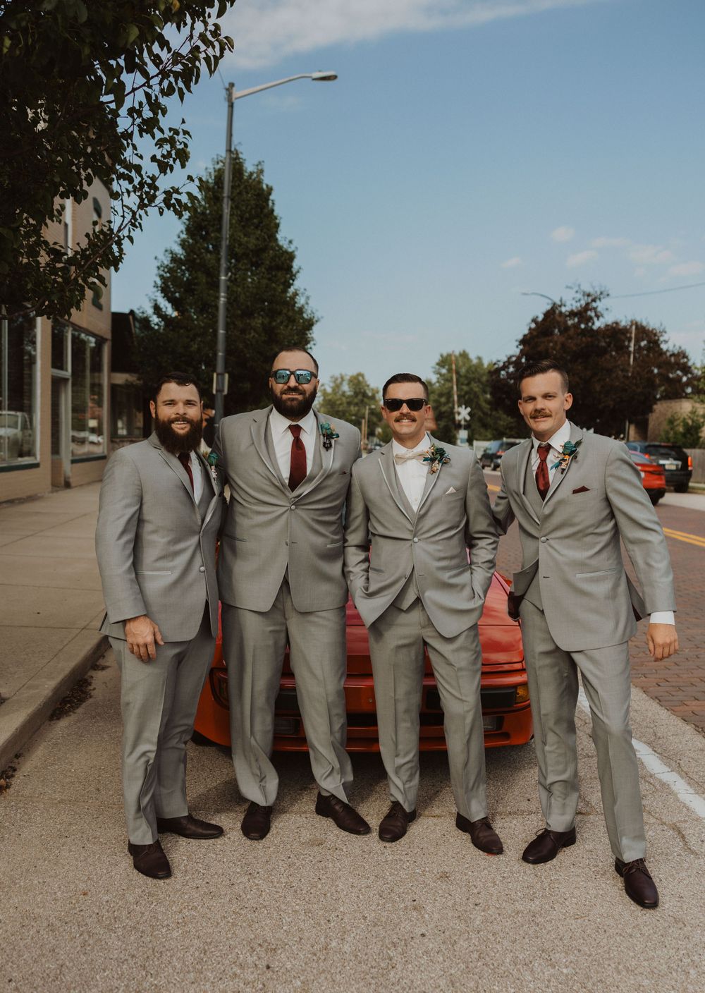 Groomsmen in matching gray suits and red ties pose together on a sidewalk next to a red car.
