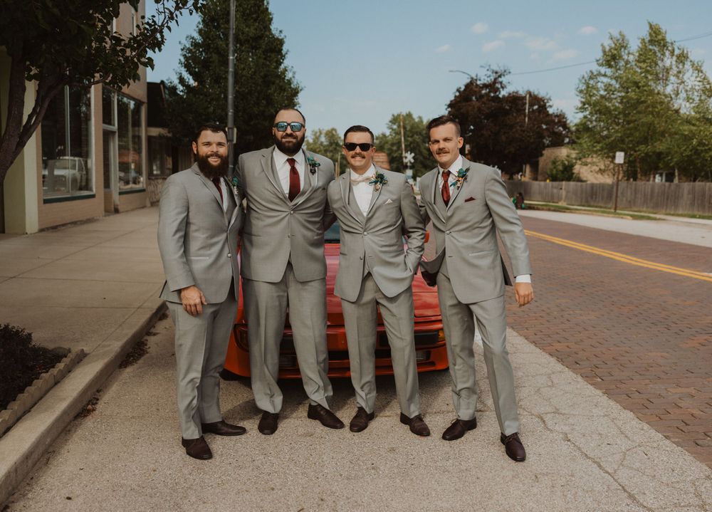 Wedding party members in gray suits sit casually on a curb during photo session.