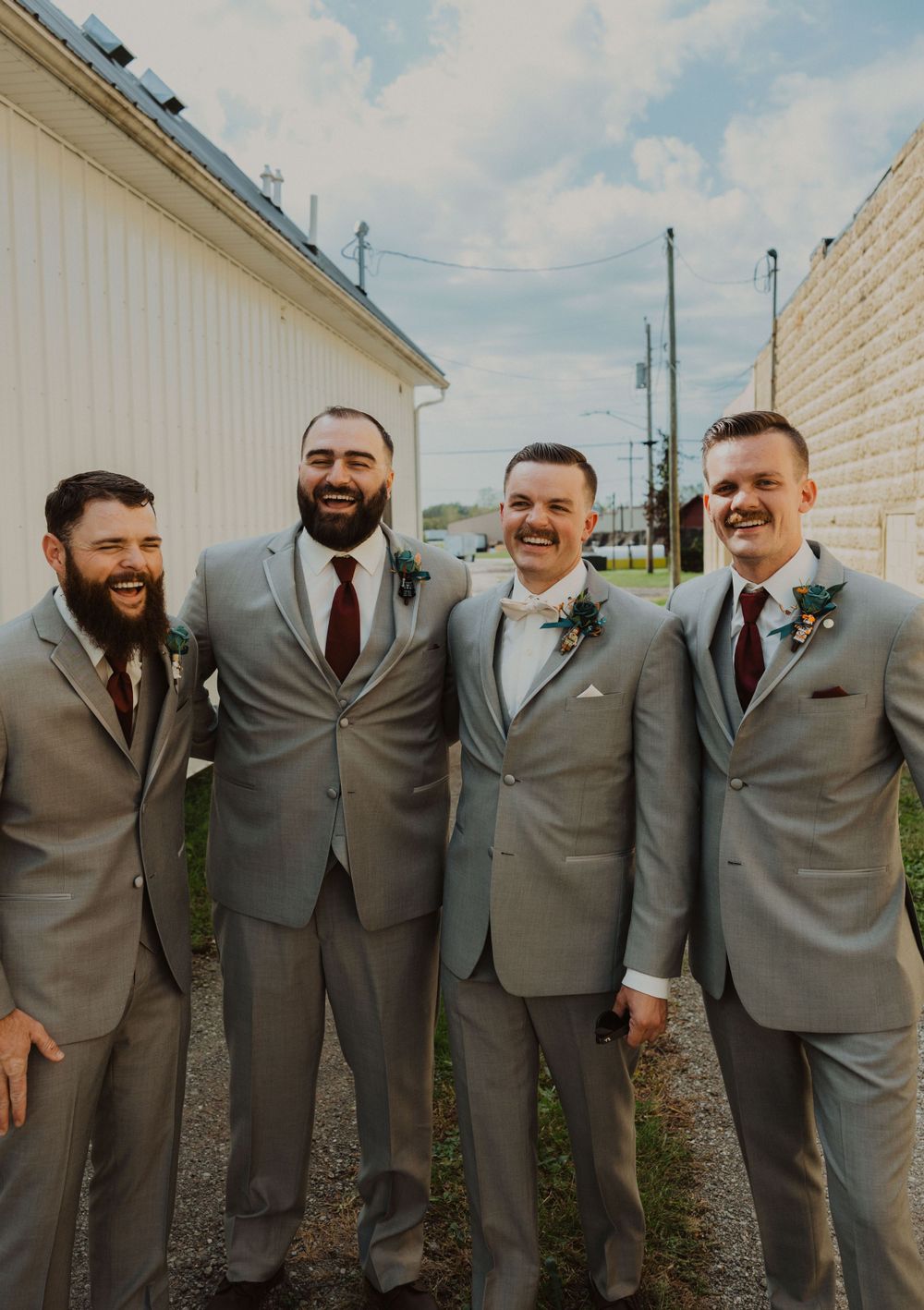 Groomsmen in coordinated gray suits and burgundy ties smile while lined up against a building.