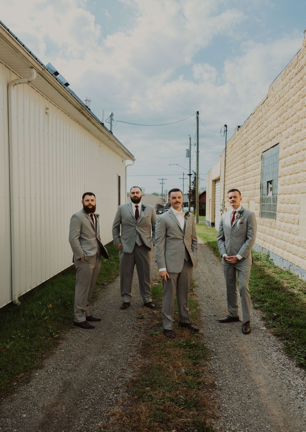 Wedding party in matching gray suits poses along a gravel path beside white buildings.