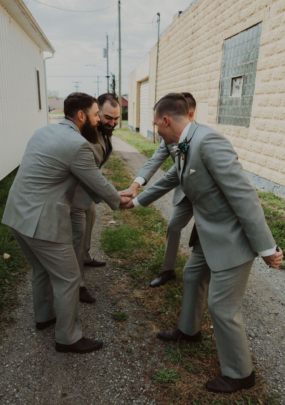 Groomsmen in gray suits playfully pretend to fight in an alleyway.