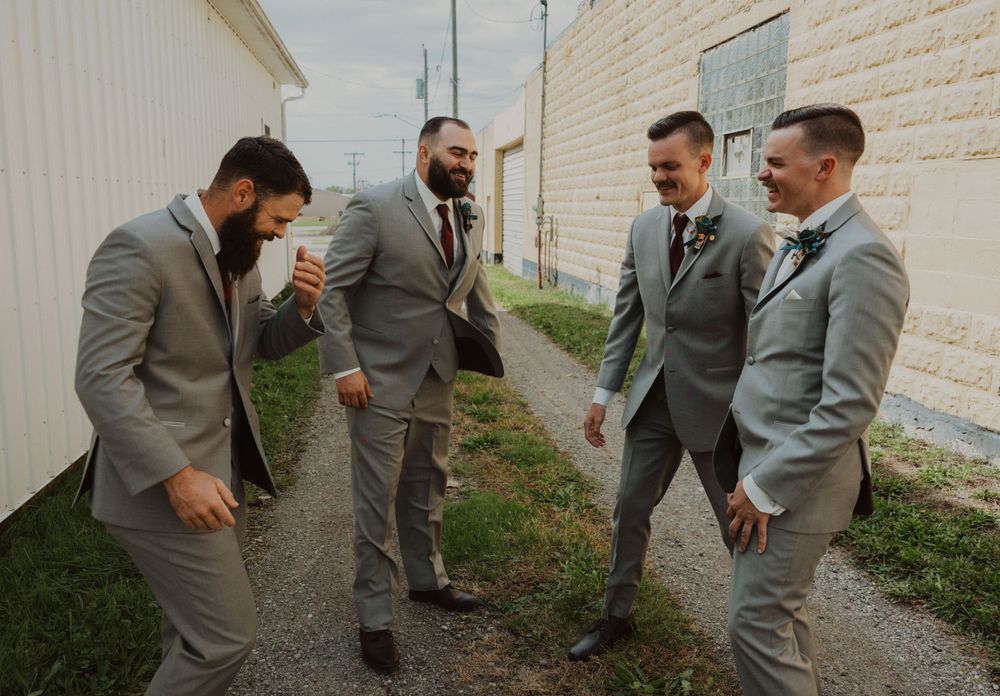 Wedding party members in gray suits engage in playful poses between buildings.