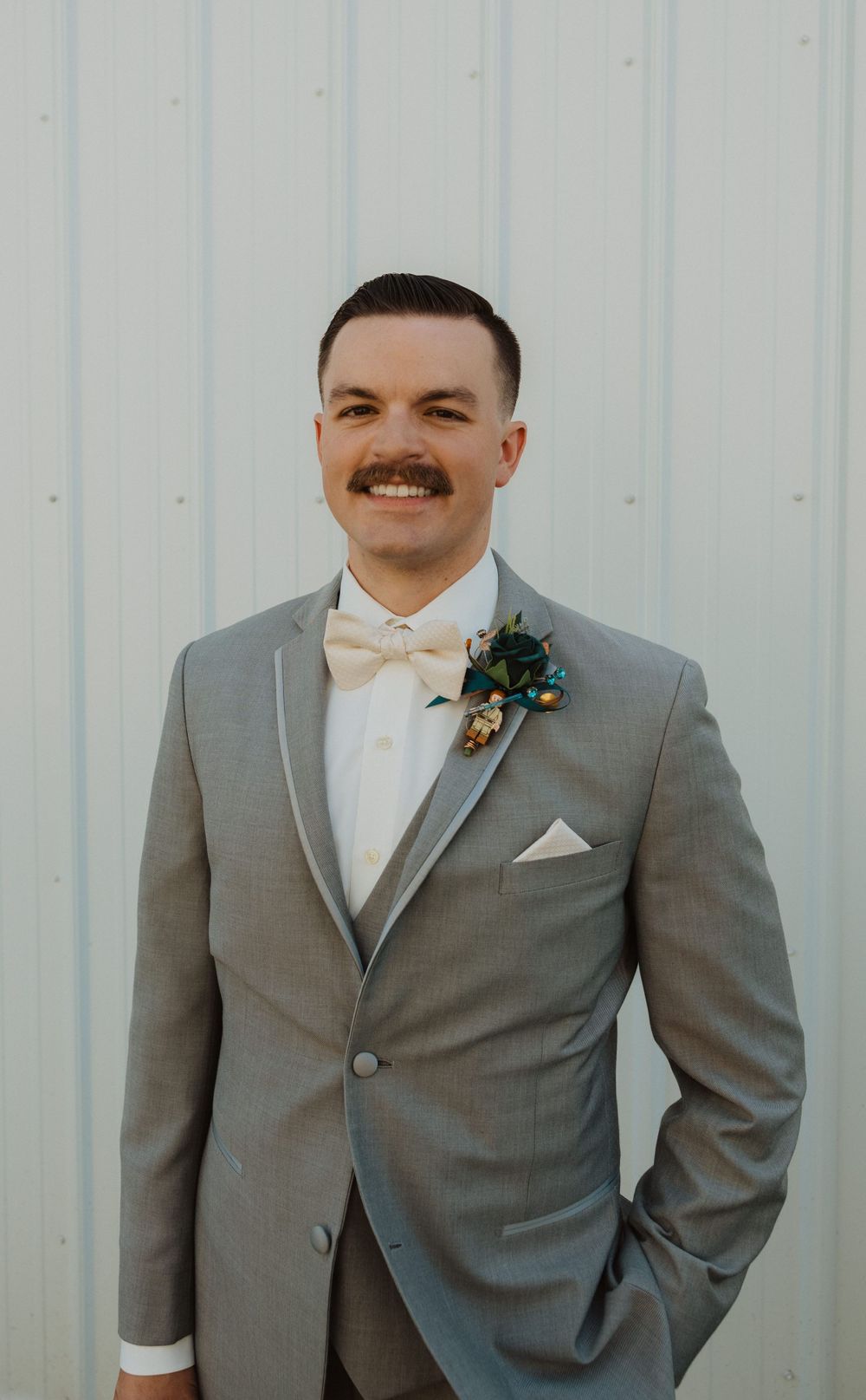 A groom in a light gray suit with bow tie and boutonniere poses against white siding.