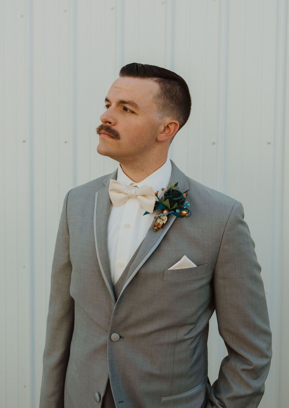 Groomsmen in gray suits with burgundy boutonnieres stand against a white wall at a wedding.