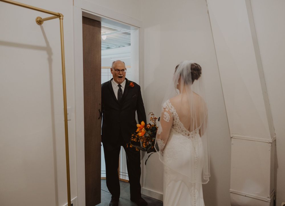 A first look moment between the bride and her father in a bright white hallway.