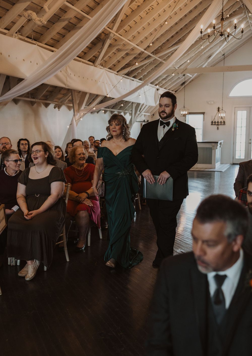 Wedding guests gather in an elegant white-walled venue with exposed wooden beams and chandeliers.