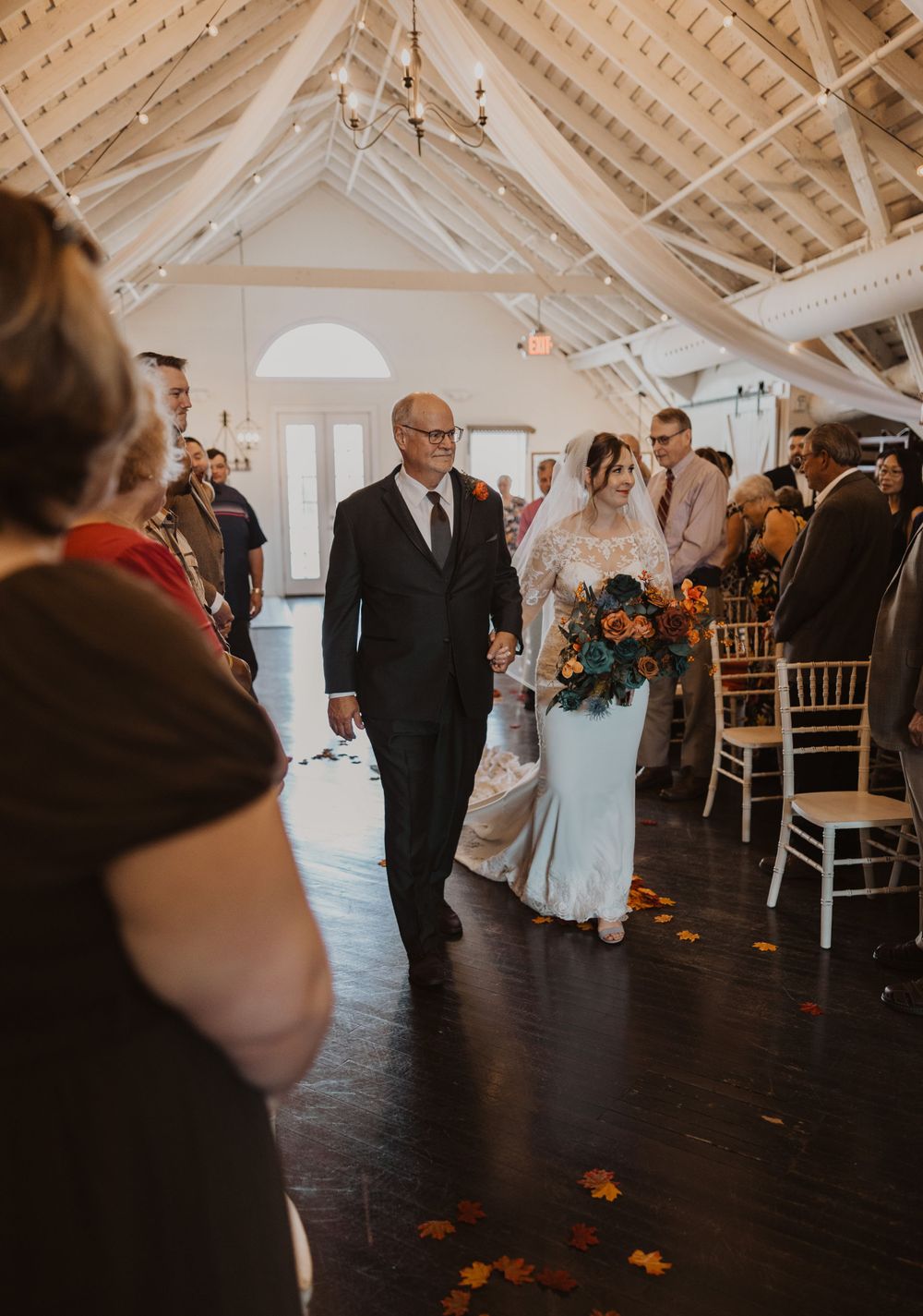 Wedding ceremony in a bright white venue with exposed beams and scattered flower petals on the floor.