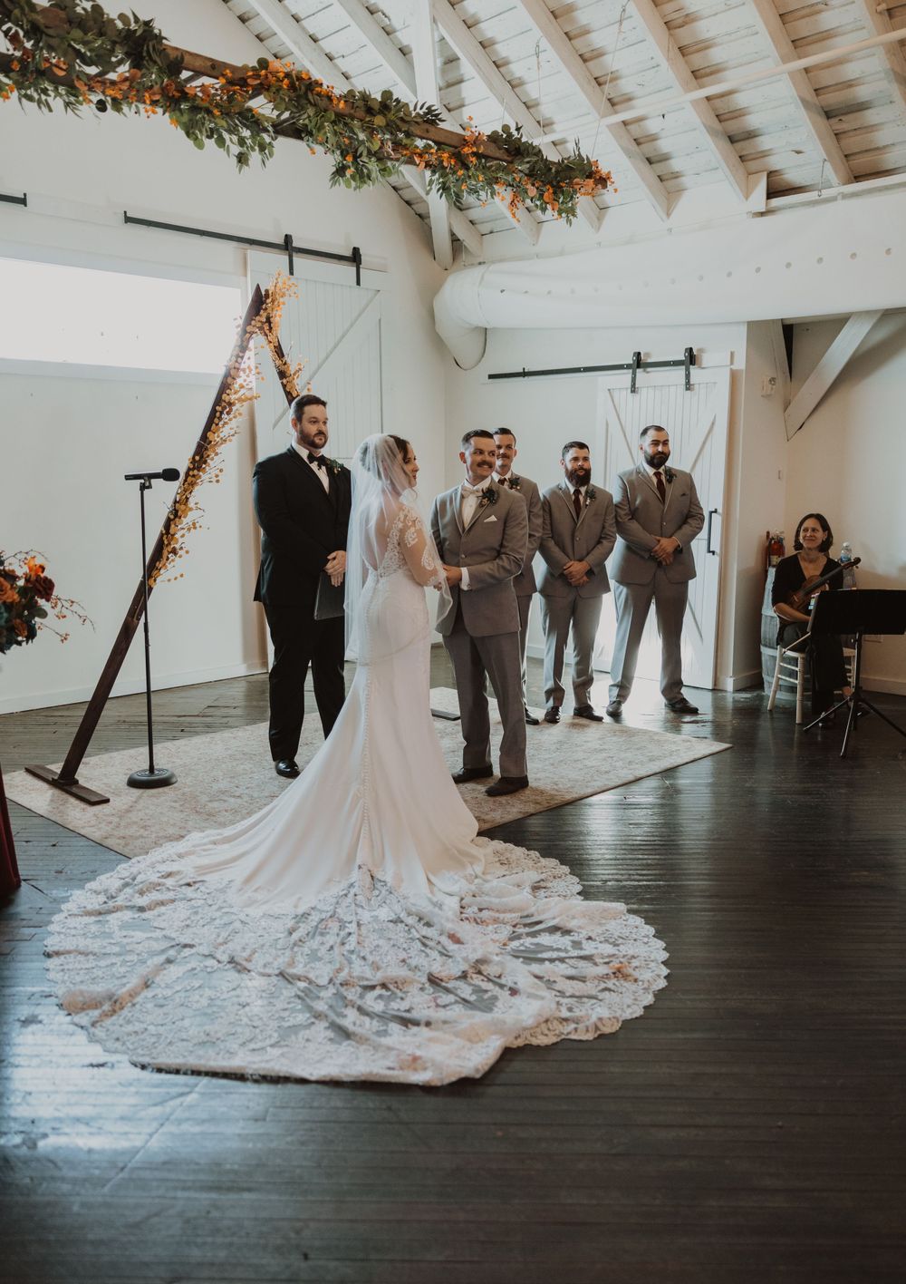 Wedding party stands in rustic white barn venue with dramatic long train wedding dress.