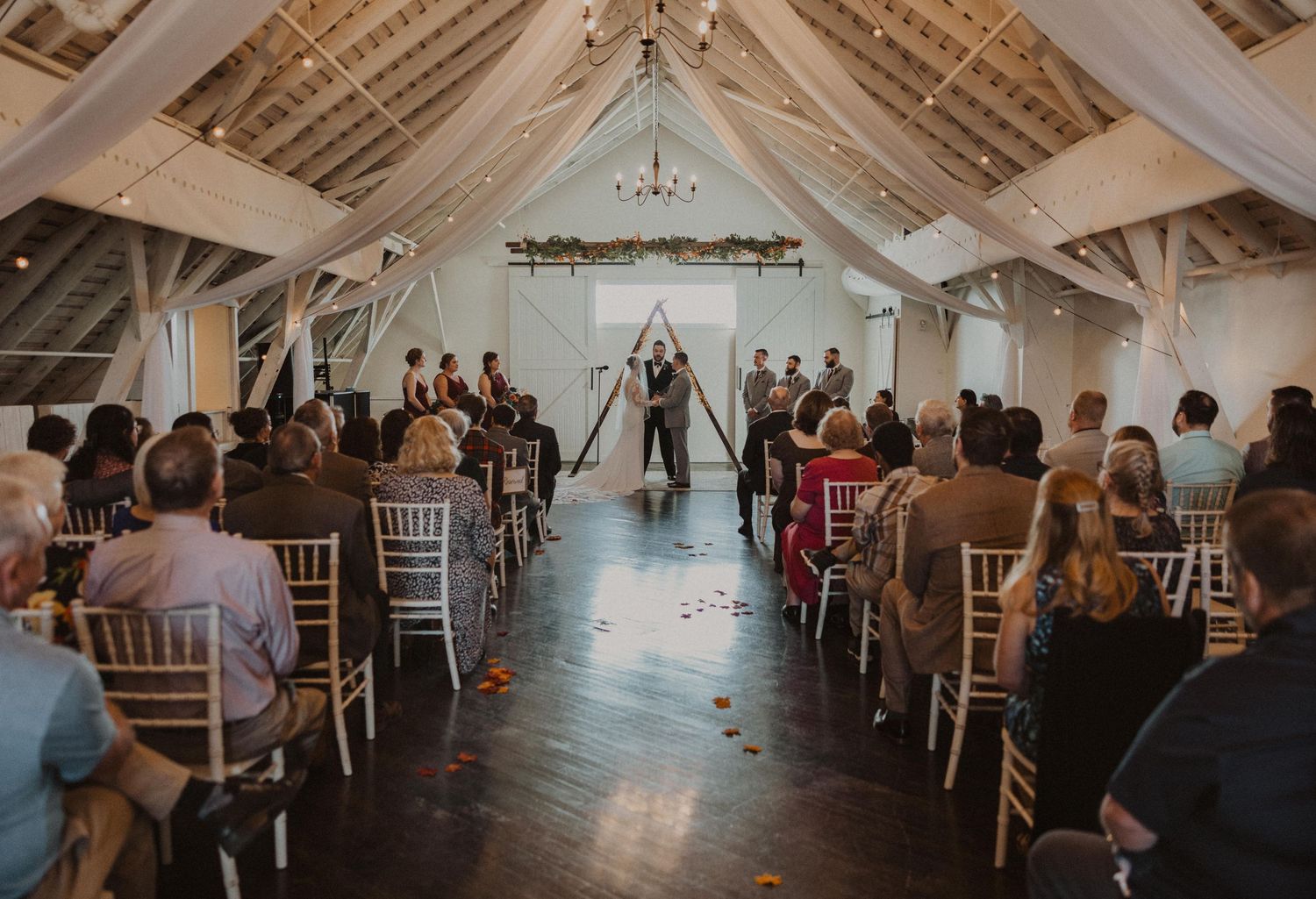 Wedding guests seated in rustic barn venue with vaulted ceilings and chandelier lighting.