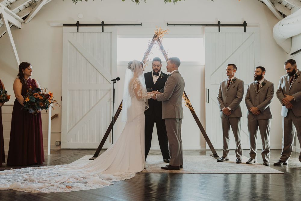 Wedding ceremony in progress inside bright white barn venue with floral ceiling decorations.
