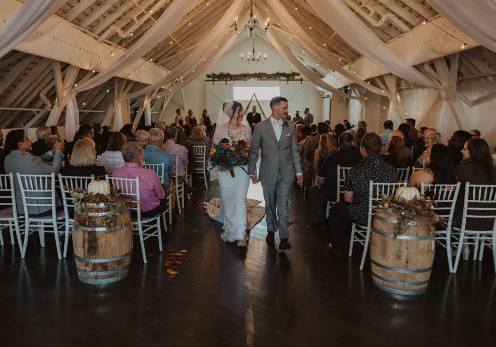 Wedding reception setup in rustic barn venue with white chiavari chairs and wine barrel decor.