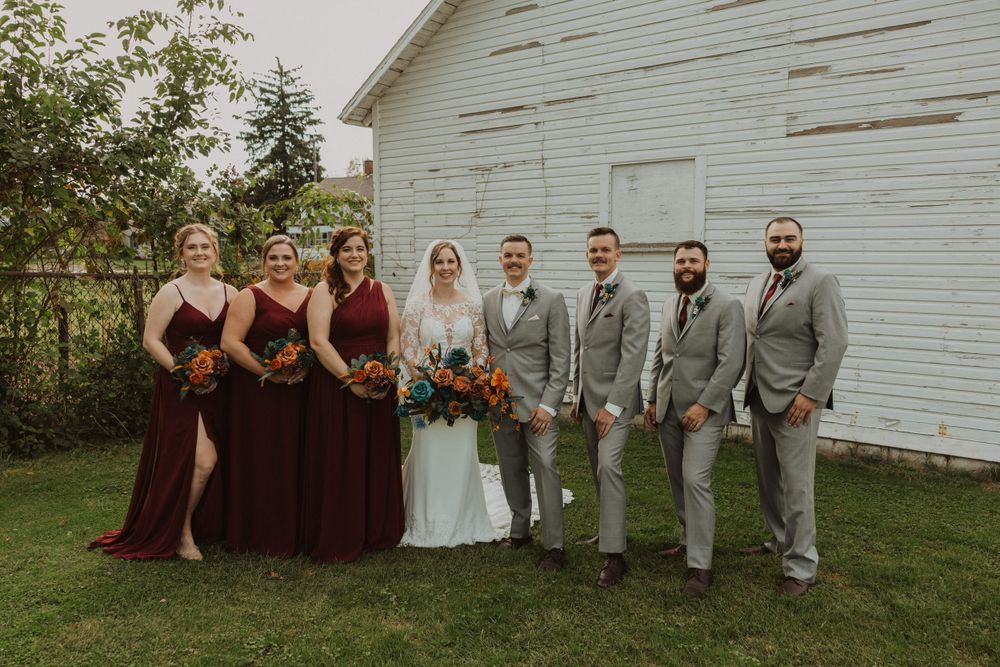 Wedding party posing in front of a white barn wearing burgundy and gray formal attire.