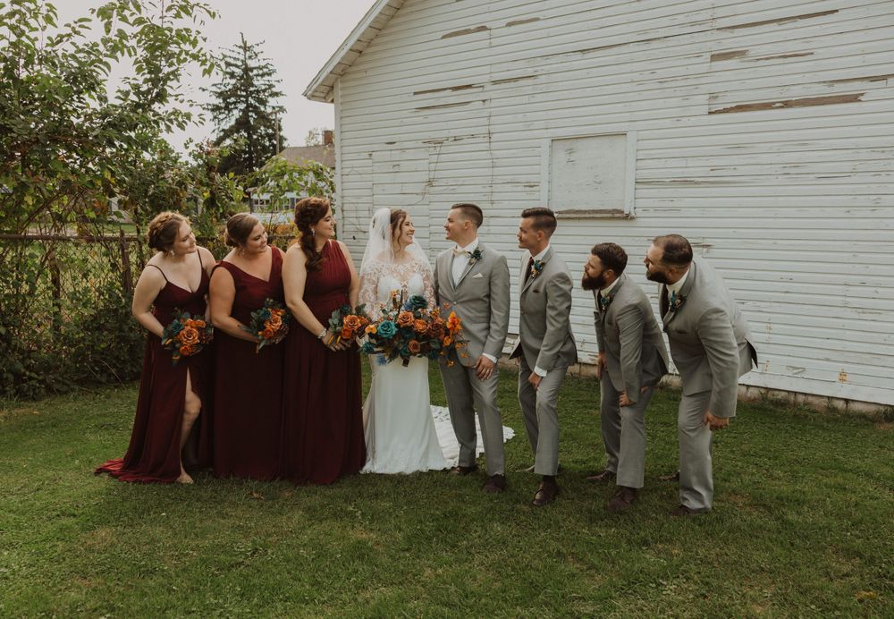 Bridal party sitting casually on grass against white barn backdrop in burgundy and gray attire.