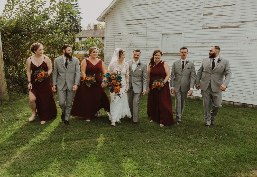 Wedding party arranged in casual poses by white barn wearing burgundy and gray formal wear.