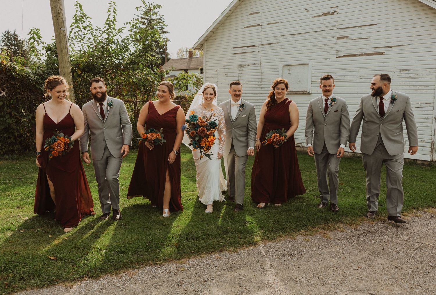 Wedding group walking and laughing together along path by rustic white barn.