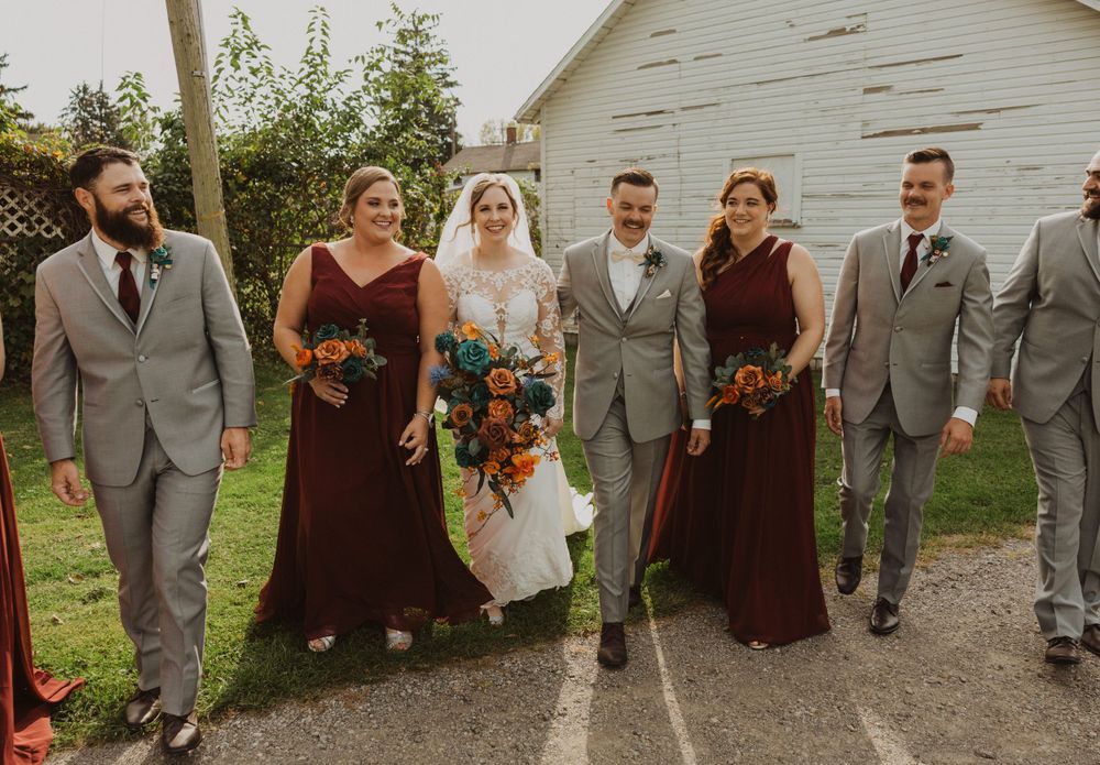 Wedding party in motion walking and jumping by white barn in burgundy and gray attire.