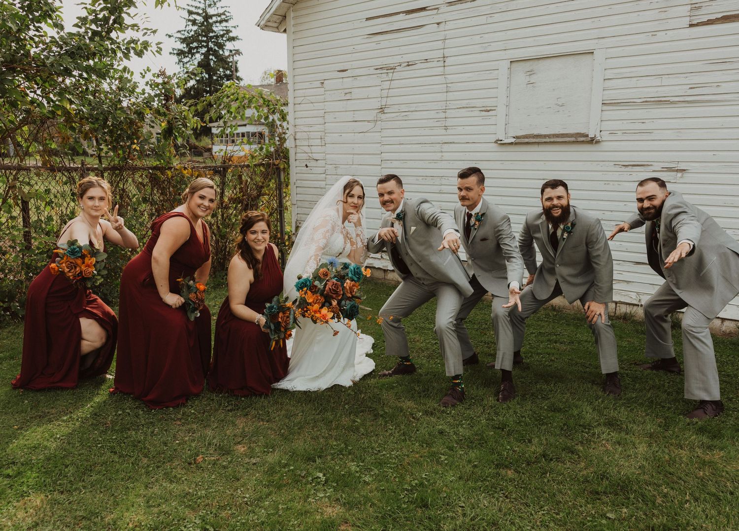 Wedding group sits casually on grass against white barn backdrop.