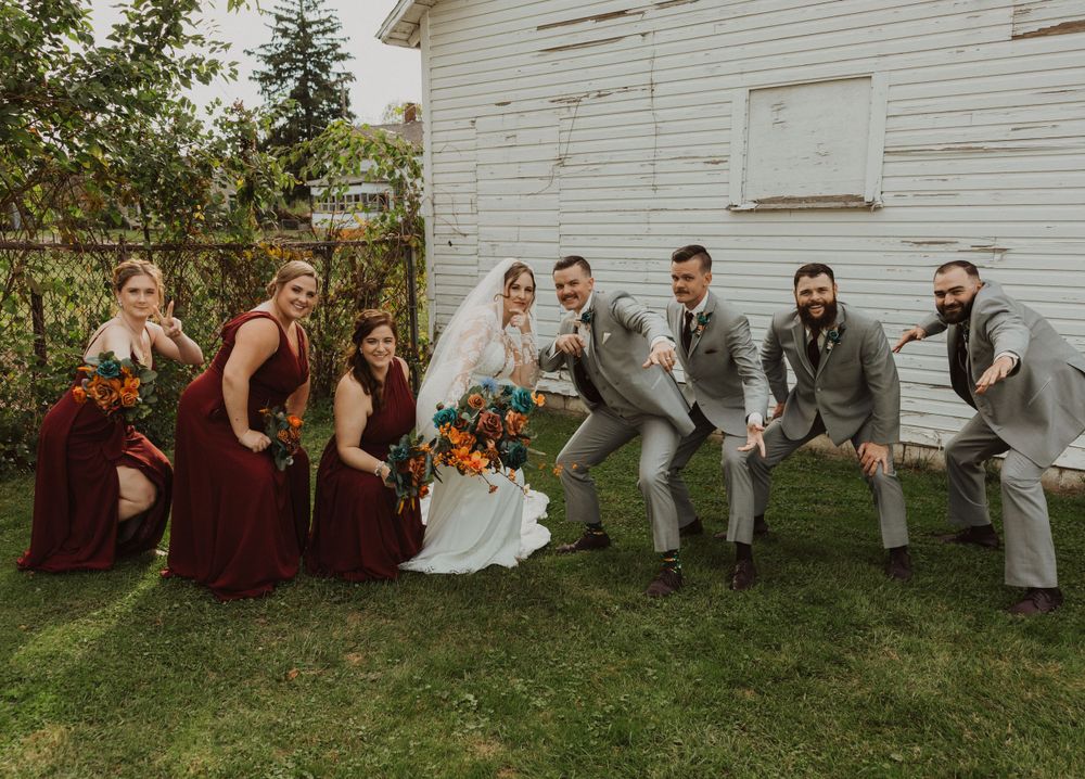 Wedding group sits casually on grass against white barn backdrop.