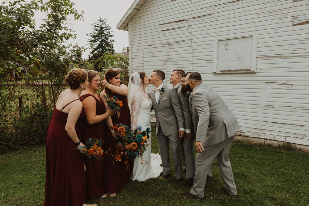 Casual wedding party poses near weathered white barn during fall celebration.