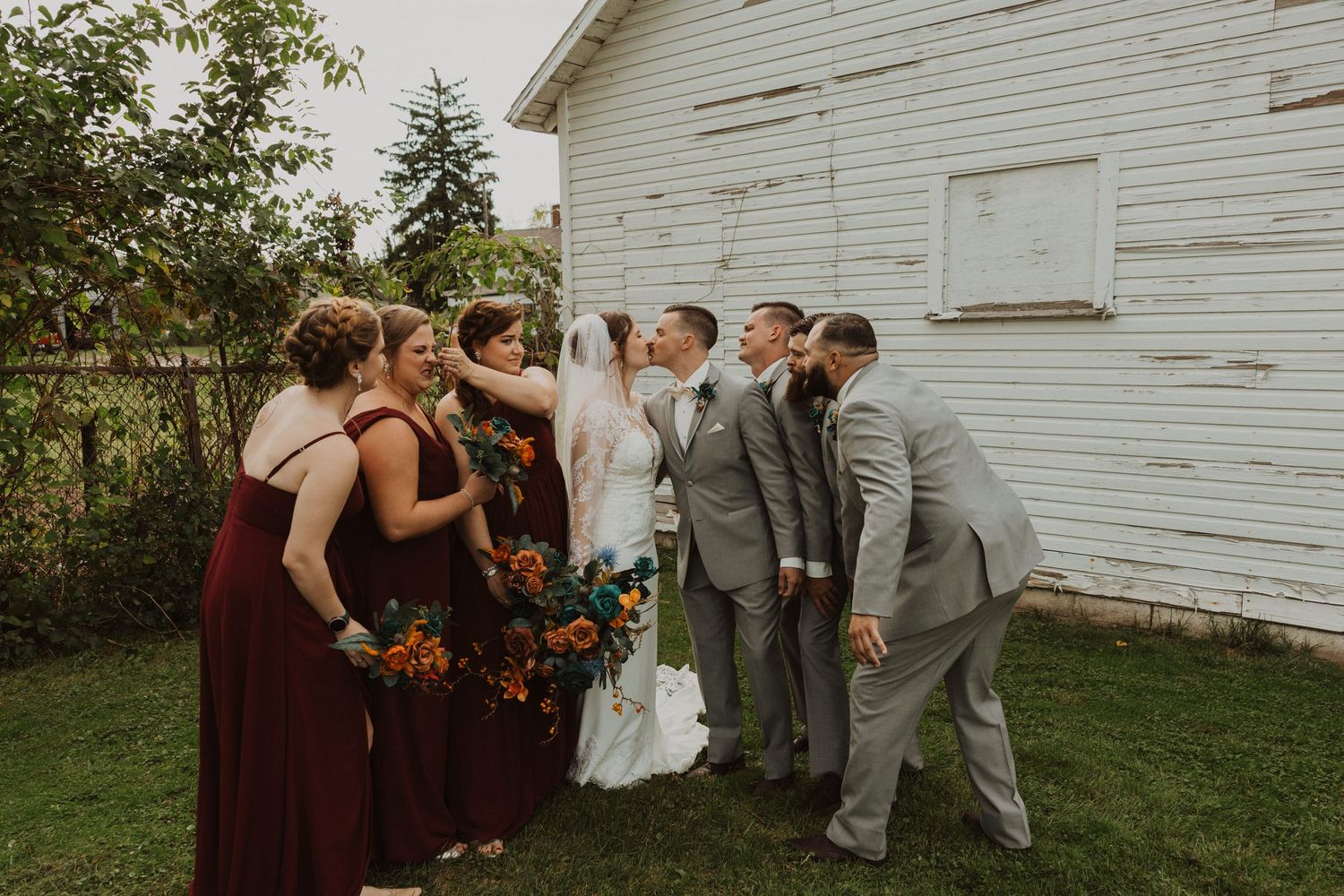 Casual wedding party poses near weathered white barn during fall celebration.