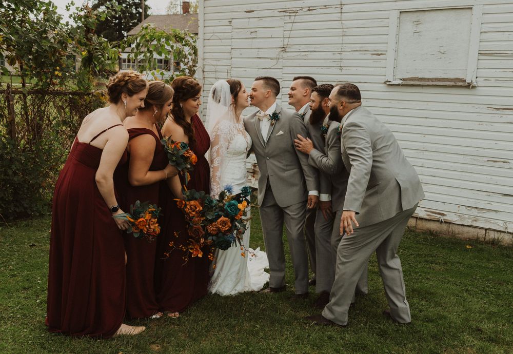Wedding party poses playfully against white barn wall with bridesmaids in burgundy and groomsmen in gray suits.