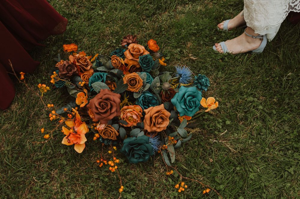 Orange and teal floral bouquets lay scattered on green grass during wedding photography.