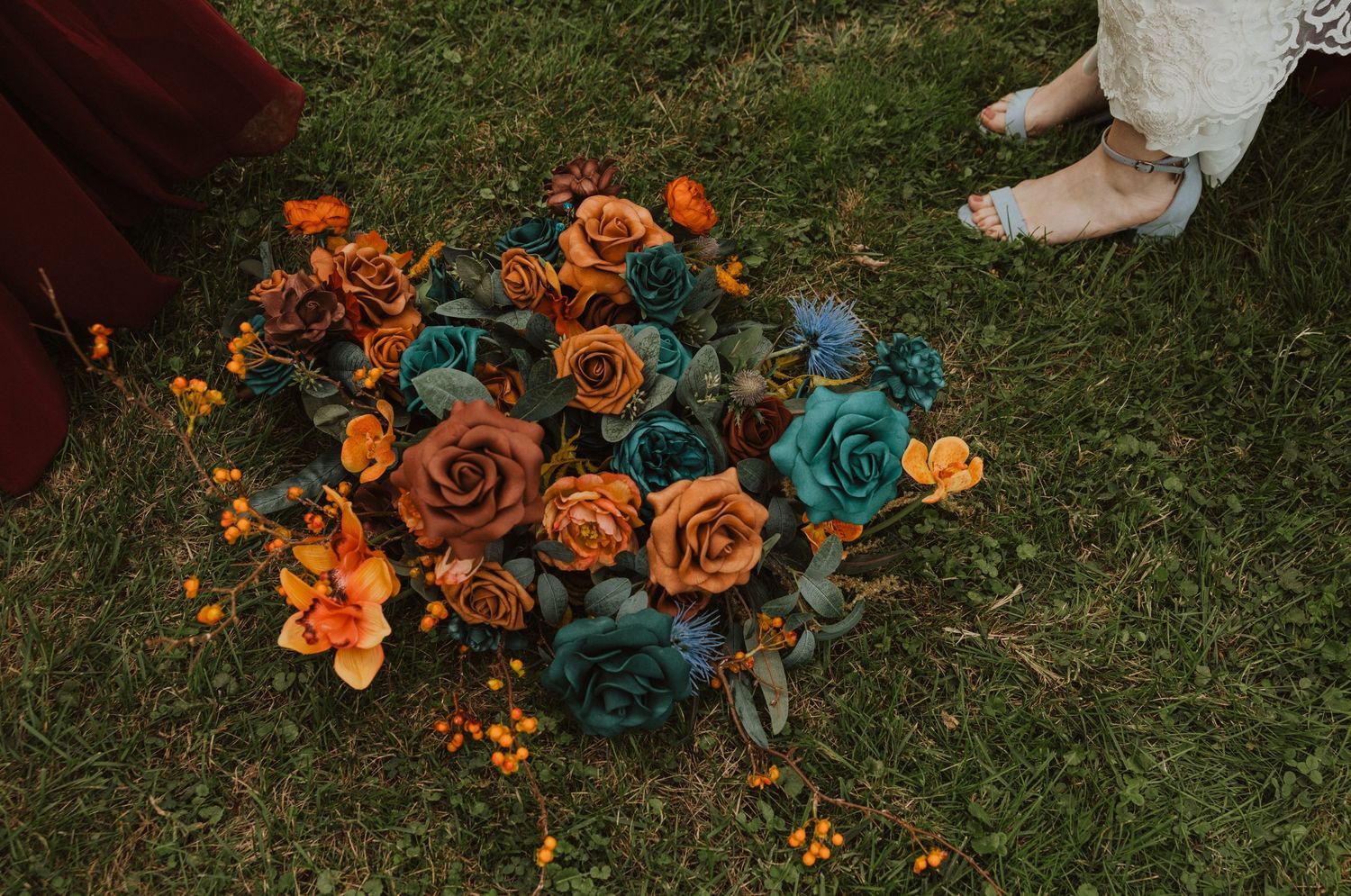 Orange and teal floral bouquets lay scattered on green grass during wedding photography.