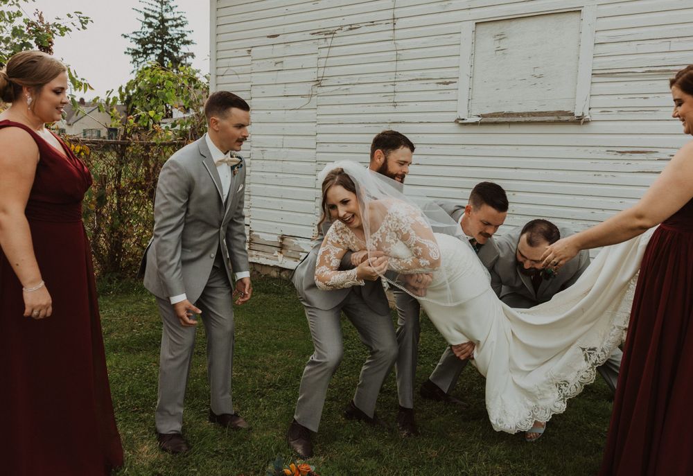 Wedding party poses playfully on lawn in front of white building with burgundy and gray attire.