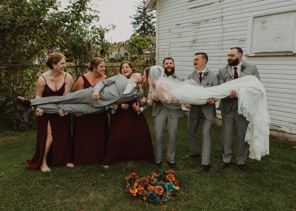 Wedding group poses playfully on grass with orange flowers and white building backdrop.