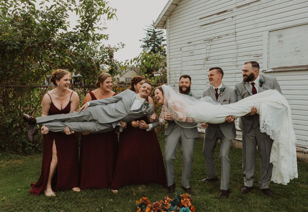 Bridal party creates domino effect pose on grass near white farmhouse.