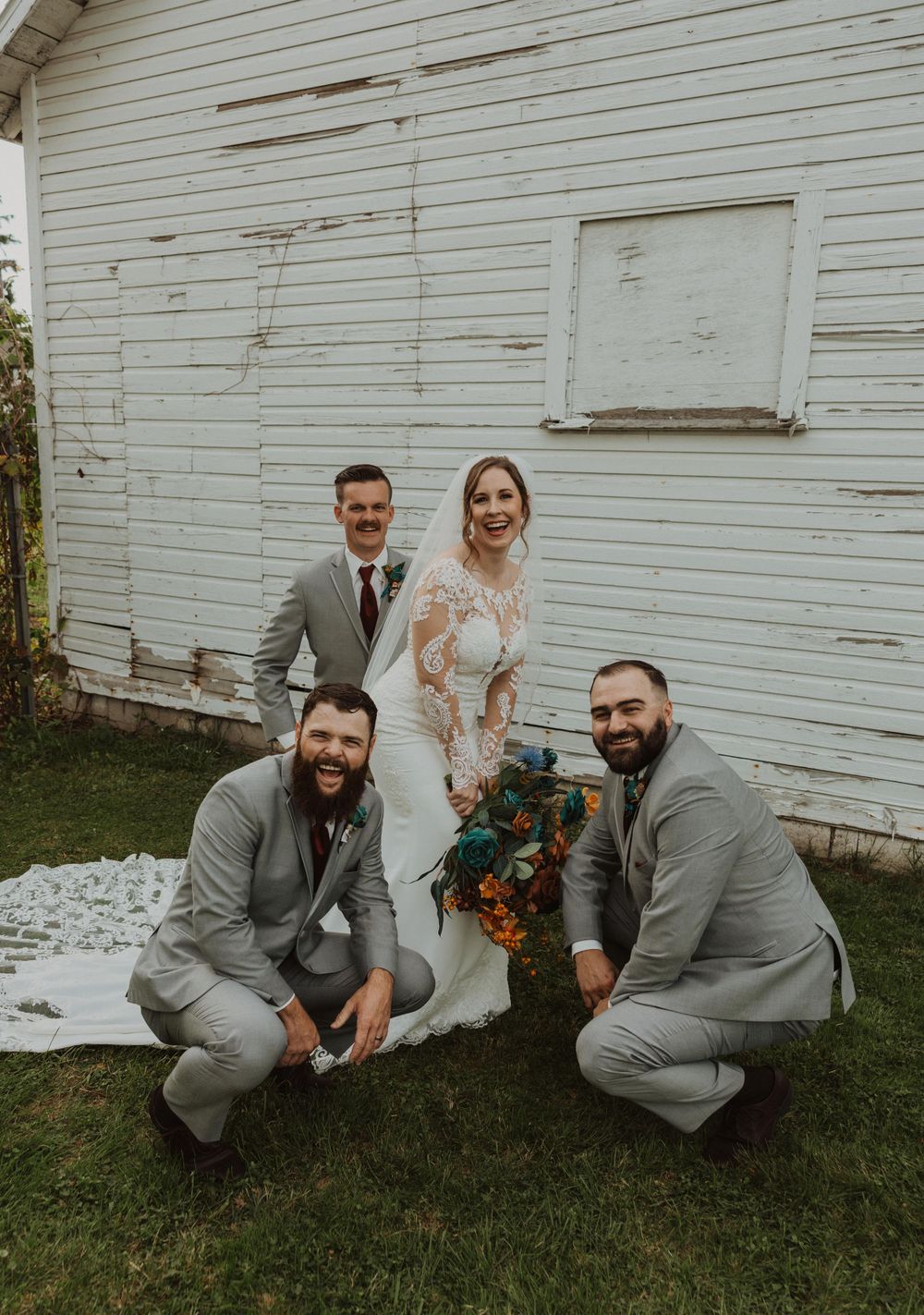 Groomsmen in gray suits crouch down by rustic white barn wall.