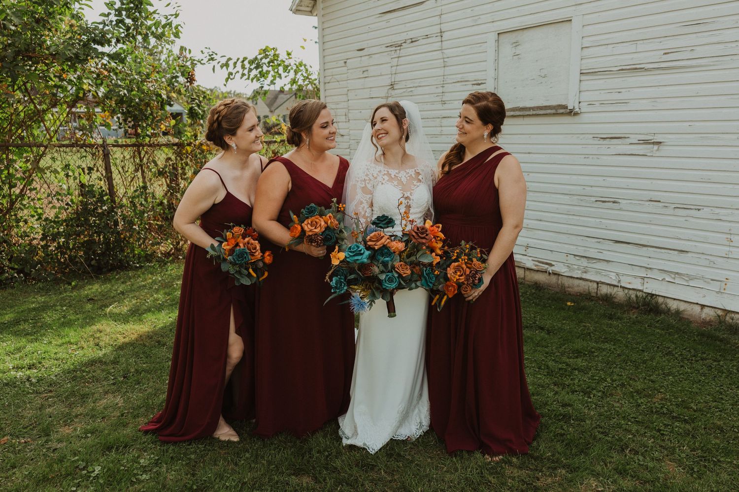 Bridal group in burgundy and white poses against weathered white building.