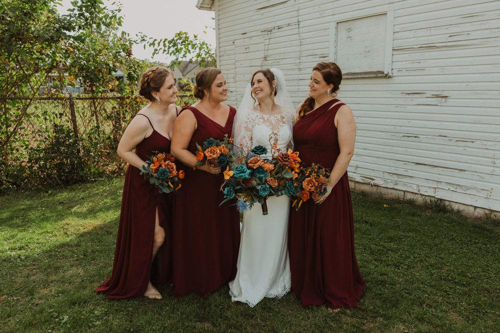 Bridal group in burgundy and white poses against weathered white building.