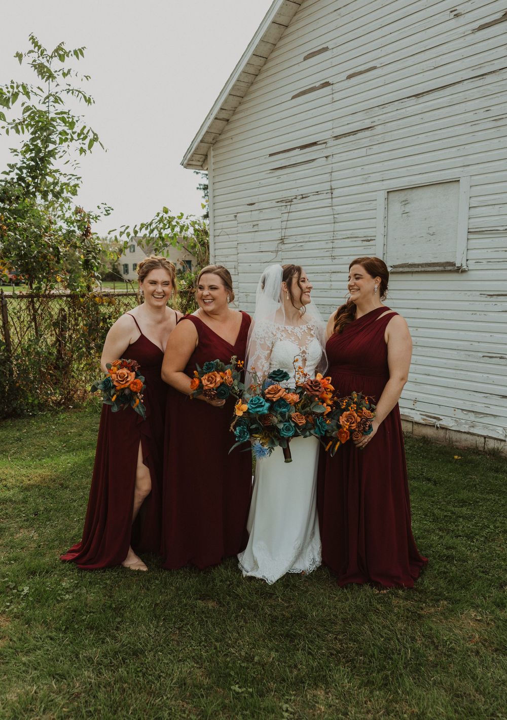 Bridal party poses casually in burgundy dresses by rustic white church.