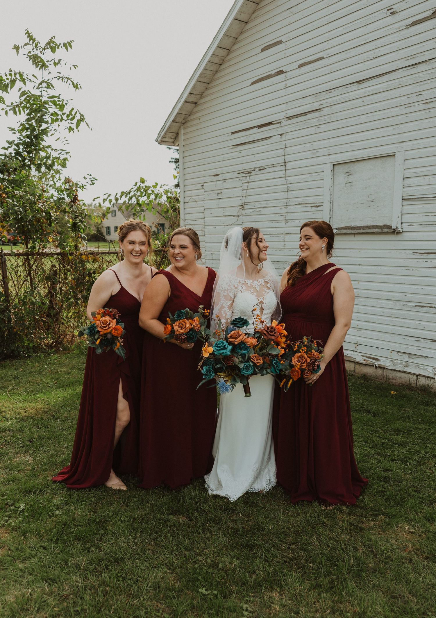 Bridal party poses casually in burgundy dresses by rustic white church.