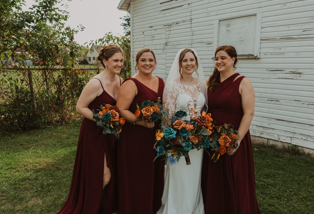 Bridesmaids in burgundy dresses share candid moment with bride in front of weathered white building.