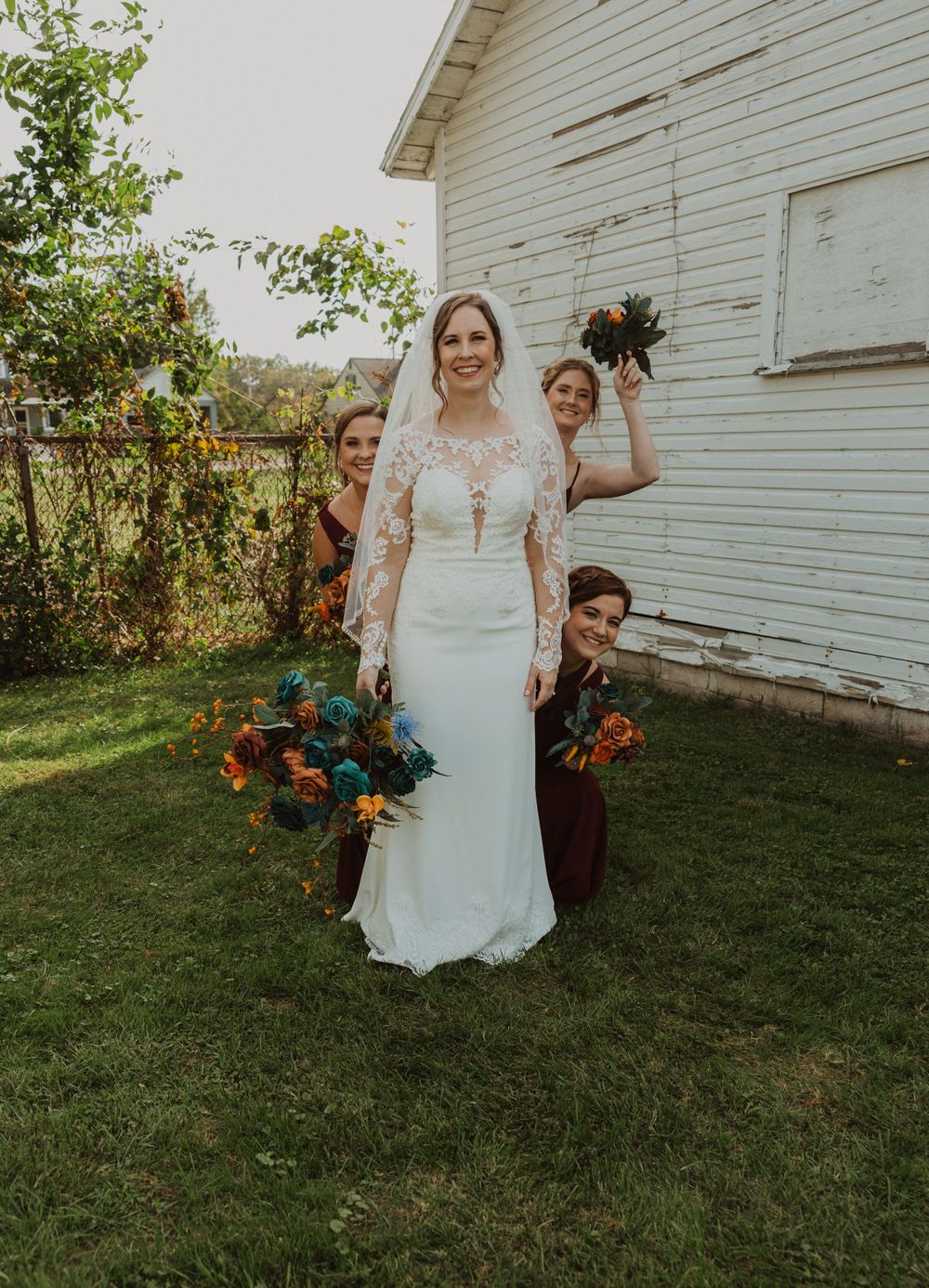 Wedding party poses playfully on grass in front of white barn with fall-colored floral bouquets.