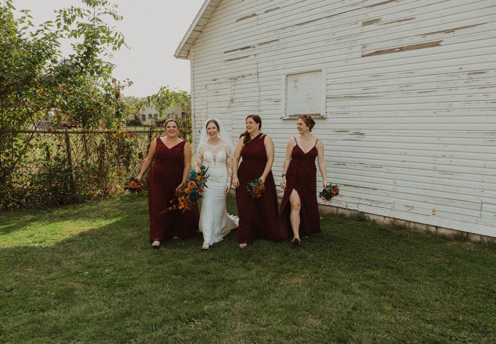 Bridal party poses informally against white barn siding in autumn outdoor setting.