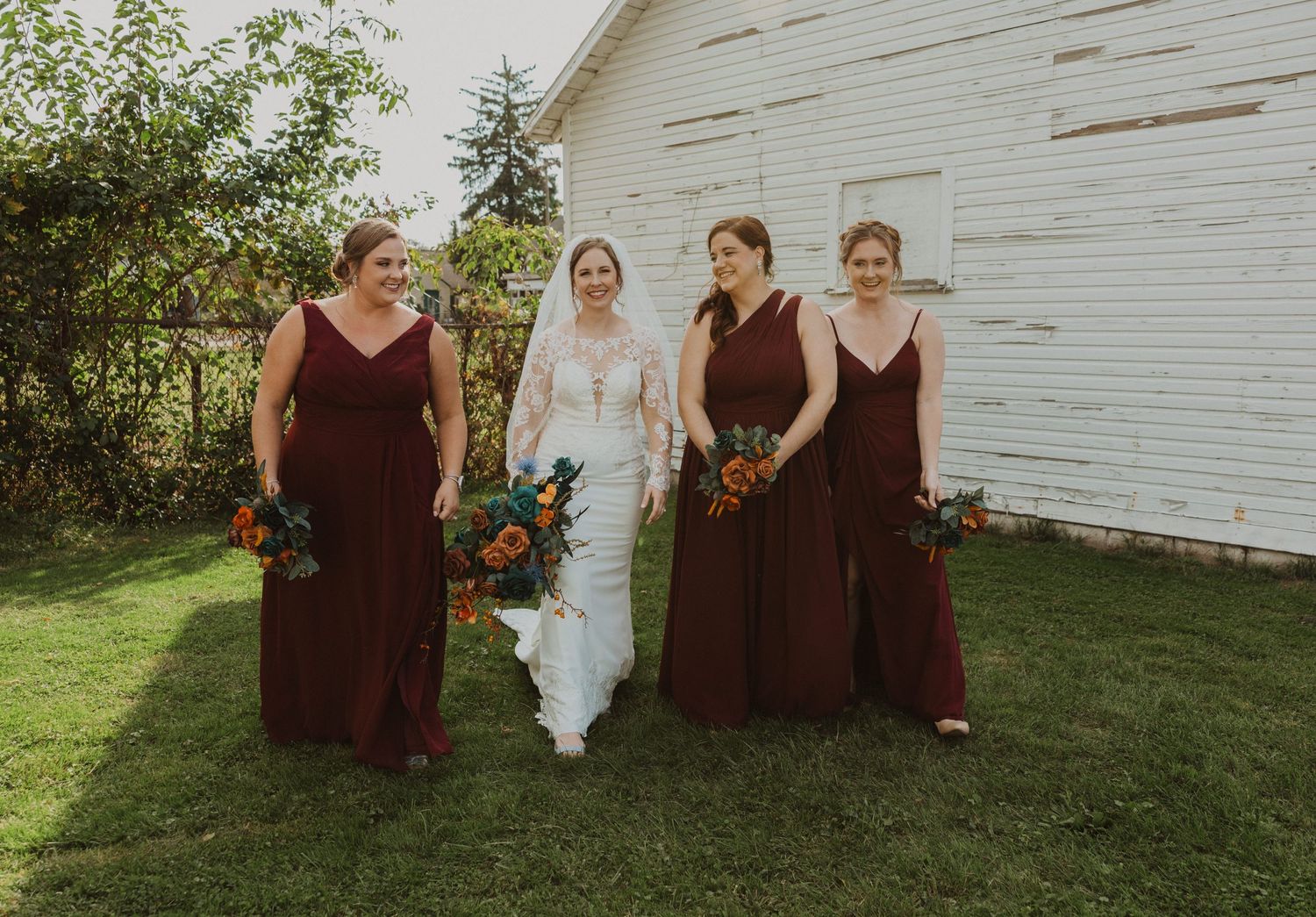 Bridal group in burgundy dresses poses informally outside white barn with fall flowers.