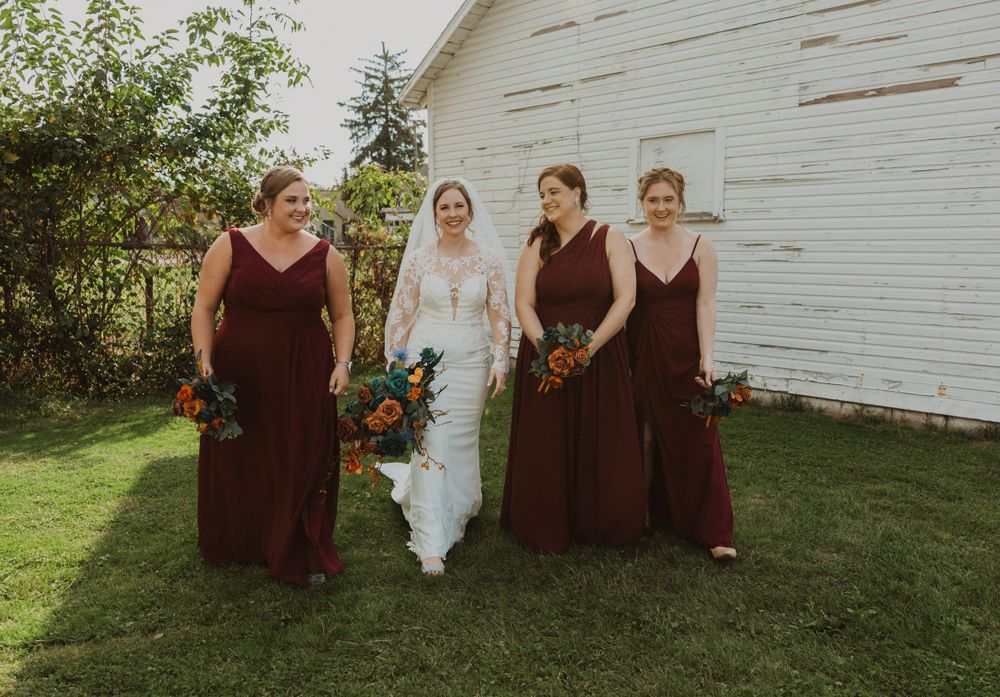 Bridal group in burgundy dresses poses informally outside white barn with fall flowers.