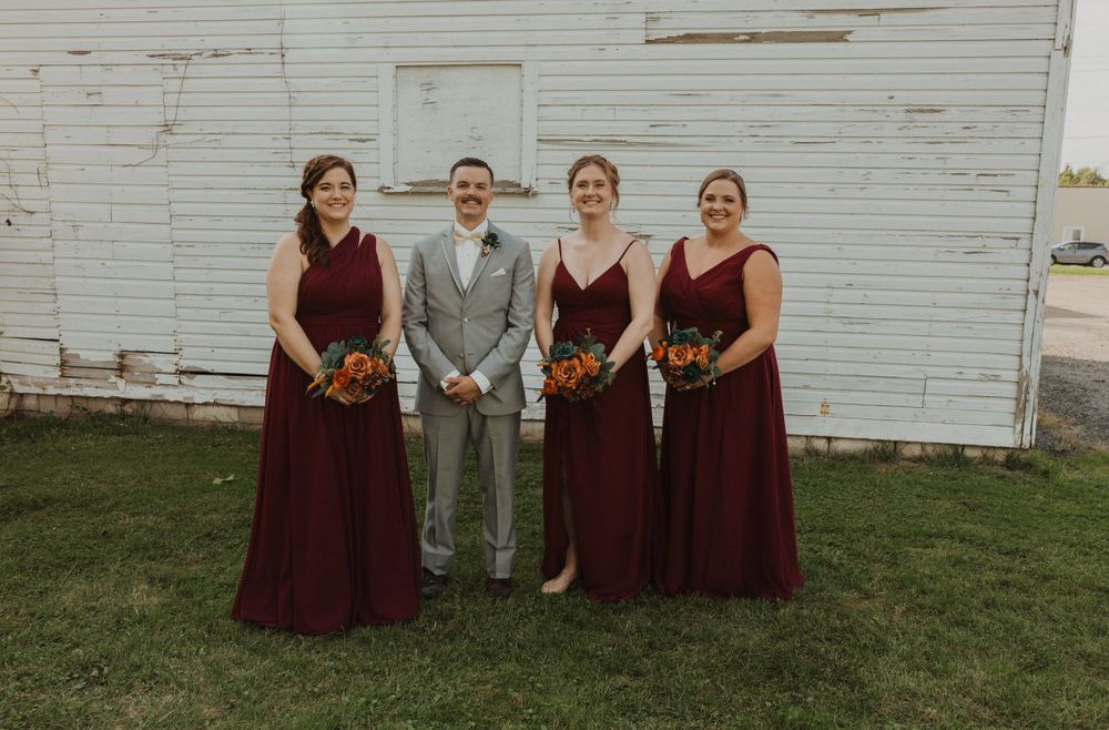 Wedding party members relax on lawn in front of weathered white barn.