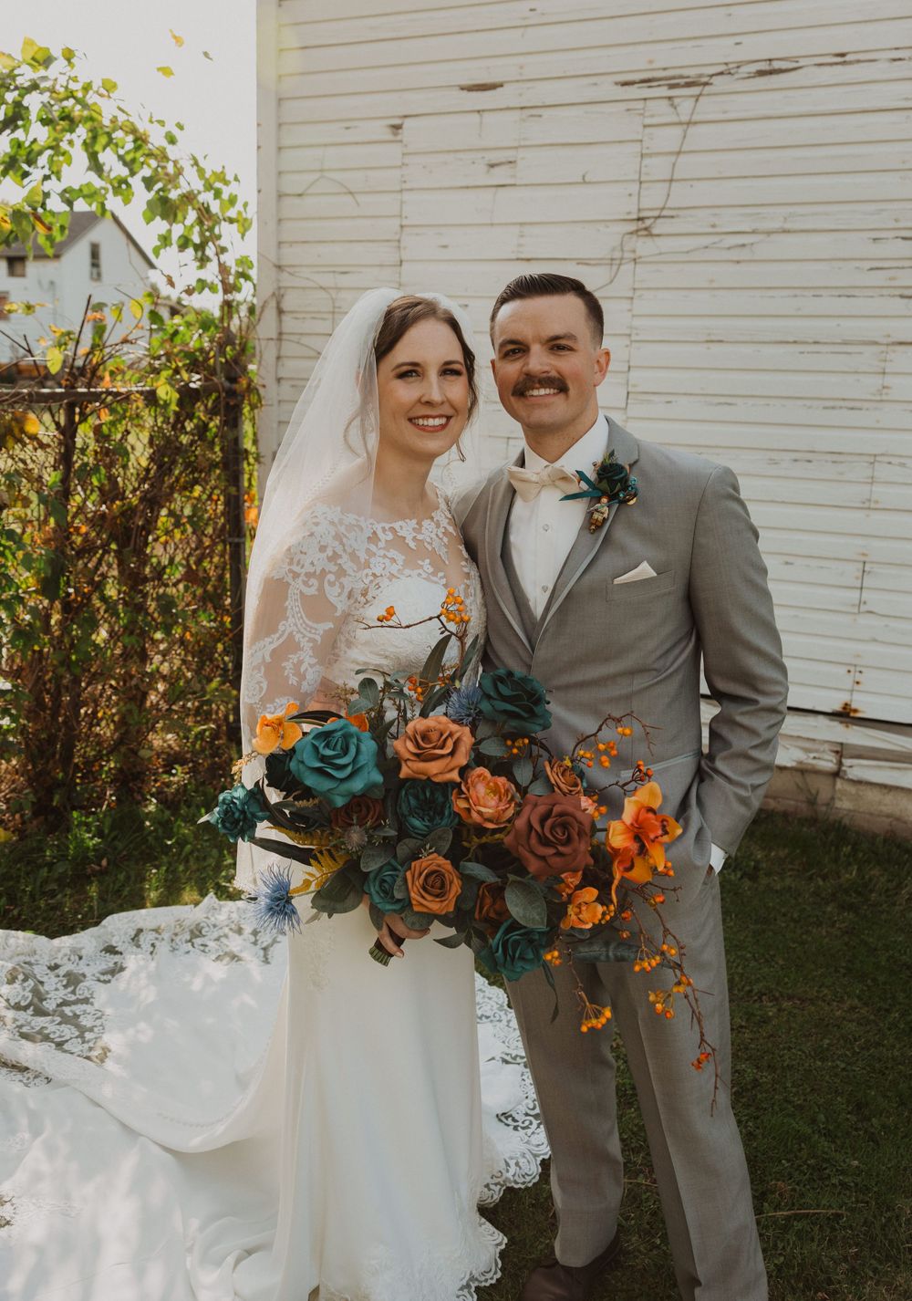 Couple poses with orange and teal bouquets against white barn on their wedding day.