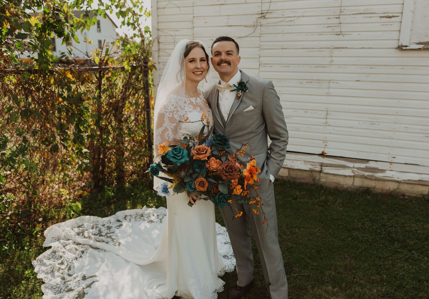 Newlyweds stand together holding vibrant fall bouquets by rustic barn.