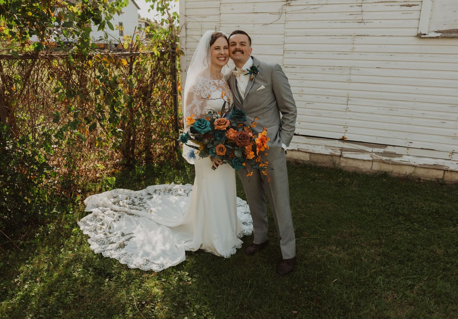 Wedding couple poses outdoors with orange and teal floral arrangements.