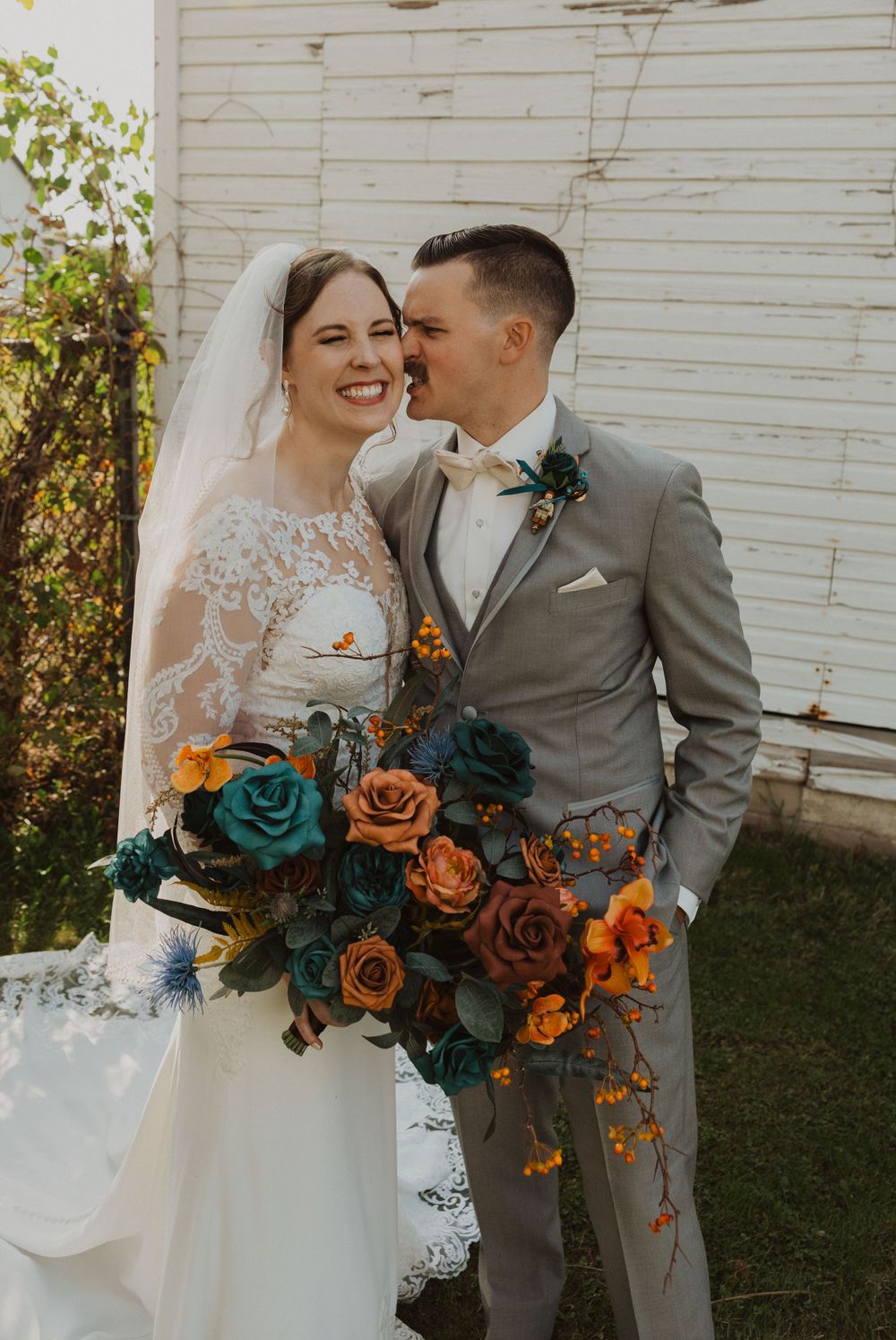 Beautiful bridal bouquet features teal and orange roses against white lace dress and gray suit.
