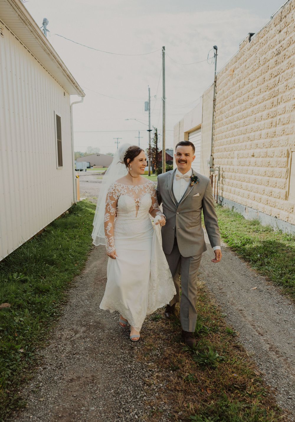 Couple strolls along gravel path between white buildings during wedding photoshoot.